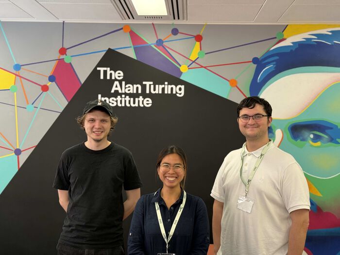 Three PhD students in front of a wall with a painting of Alan Turing.