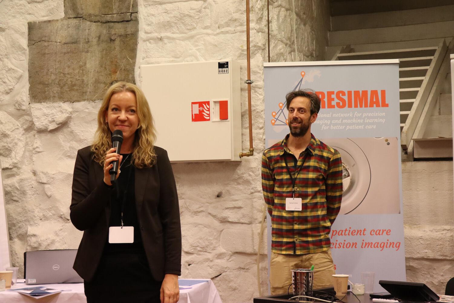 Two people presenting, a PRESIMAL poster in the background