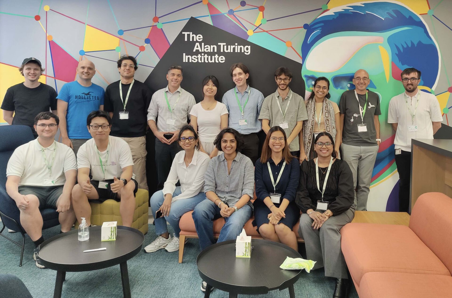 16 people in front of a recognizable wall with a painting of Alan Turing at the ATI Institute.