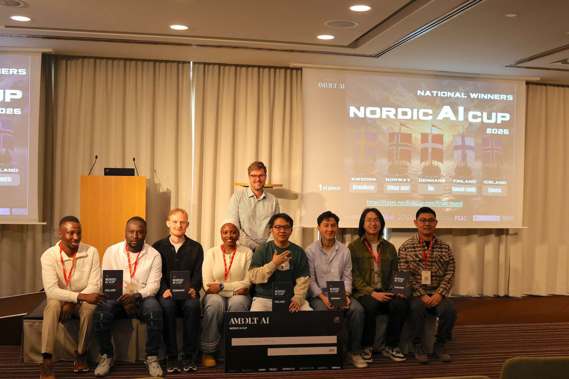 Eight people sitting on stage, one person on his knees behind them. The text "Nordic AI Cup" visible on a screen.