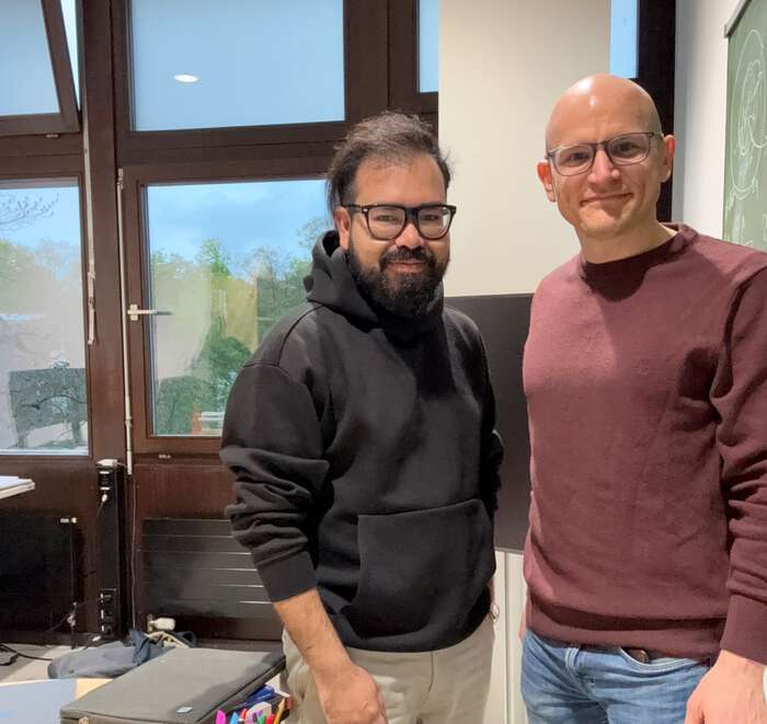 Two men with glasses standing next to each other. One with black hair and sweatshirt, one bald with a dark read sweater.