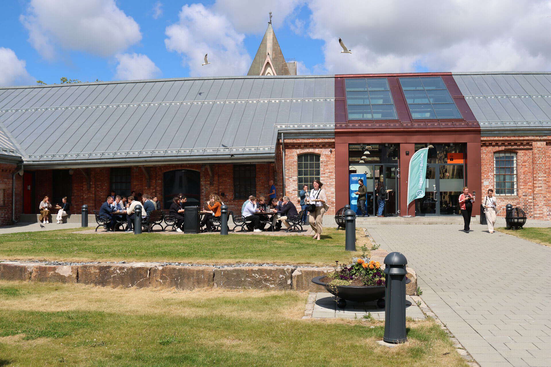 Lav mursteinsbygning, gress, folk som sitter utenfor og spiser
