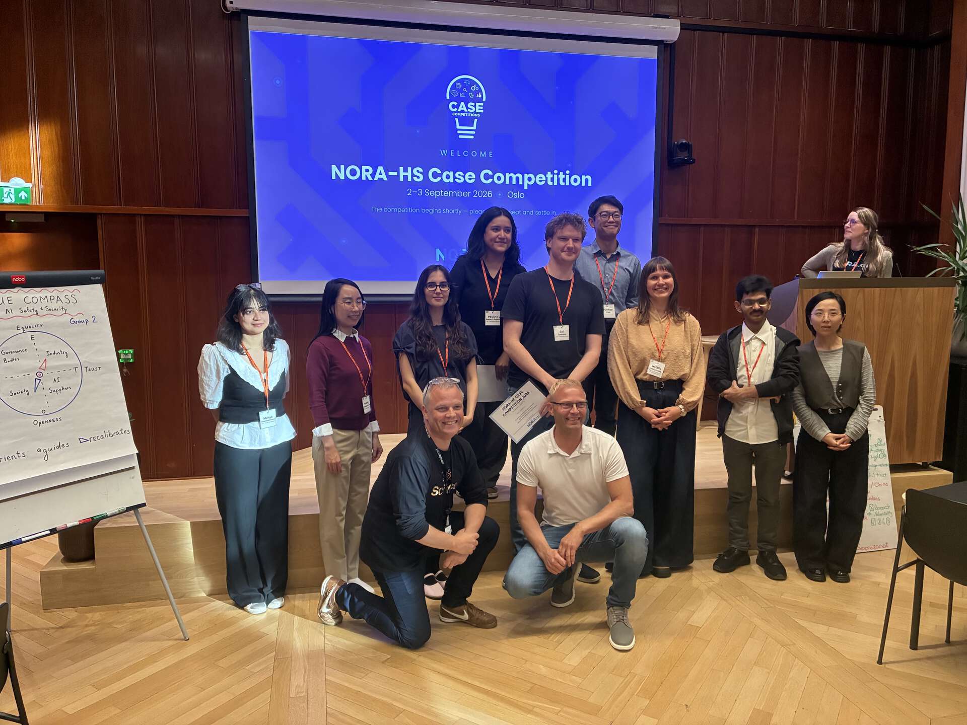 NORA-HS members and case developers standing and sitting in front of a screen, wooden floors
