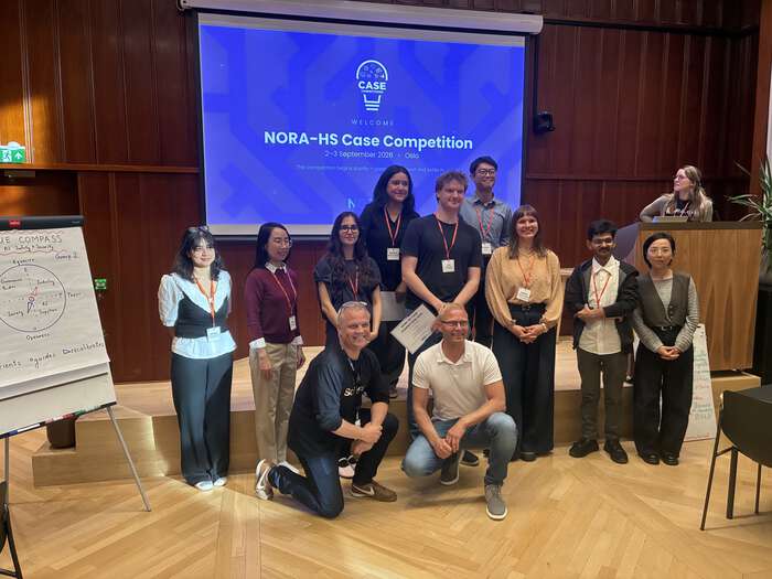 NORA-HS members and case developers standing and sitting in front of a screen, wooden floors