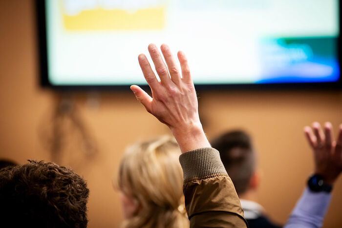 Photo of a raised hand in a room full of people, picture taken from the back and a screen visible in front of of the room